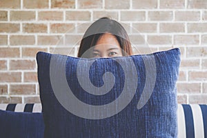 A beautiful Asian woman sitting on sofa and playing with a big blue pillow to cover her face with feeling happy and relax in cafe