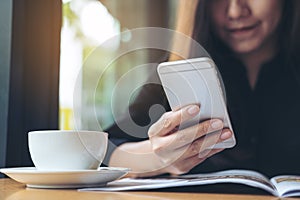 A beautiful Asian woman with smiley face holding and using smart phone while reading magazines with coffee cup on wooden table