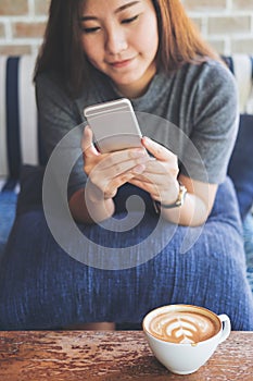 Asian woman with smiley face using and looking at smart phone sitting on sofa with white latte coffee cup on wooden table