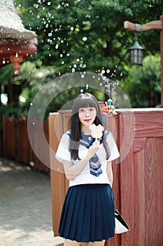 A beautiful Asian woman in student dress playing in an amusement park, eating ice cream with blow bubbles at background