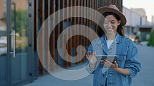 Beautiful asian woman using digital tablet, walking near the business centre texting, scrolling apps wearing brown hat