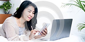 Beautiful Asian woman using smartphone and laptop  lying down in bed, female connected with technology internet