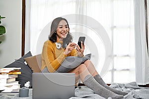 Beautiful Asian woman using smartphone, texting with her friends while relaxing in living room
