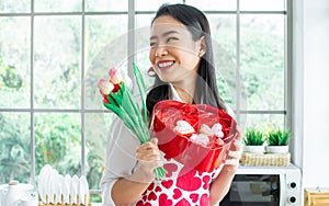 Beautiful Asian woman wearing apron with heart pattern, smiling with happiness, holding flowers, gift box of cookies, standing in