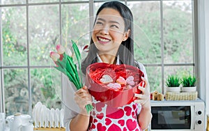 Beautiful Asian woman wearing apron with heart pattern, smiling with happiness, holding flowers, gift box of cookies, standing in