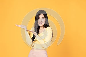 Beautiful Asian woman wearing casual clothes smile and pointing finger to an empty palm isolated on yellow background studio