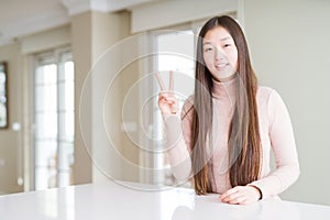 Beautiful Asian woman wearing casual sweater on white table showing and pointing up with fingers number two while smiling