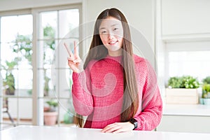 Beautiful Asian woman wearing pink sweater on white table showing and pointing up with fingers number two while smiling confident