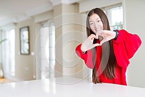Beautiful Asian woman wearing red sweater on white table smiling in love showing heart symbol and shape with hands