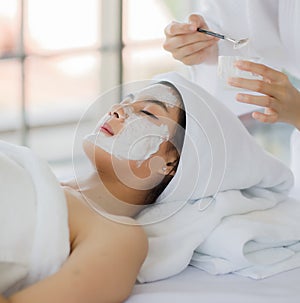 Beautiful asian woman wearing spa style white robe doing brush a facial mask herb white cream to on customer`s face for well face