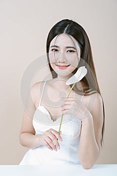 Beautiful Asian woman in white dress holding Anthurium flower on beige background. Skin care beauty treatments concept. White