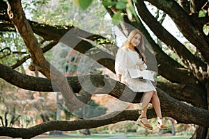 Beautiful asian woman in white dress sit on tree branch writing and thinking in the park with green leaves