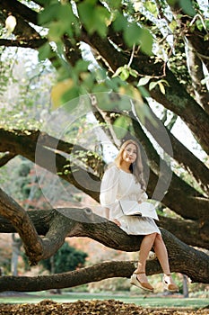 Beautiful asian woman in white dress sit on tree branch writing and thinking in the park with green leaves