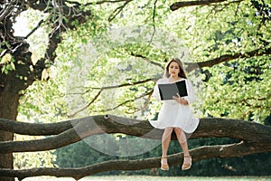 Beautiful asian woman in white dress sit on tree branch writing and thinking in the park with green leaves