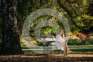 Beautiful asian woman in white dress sit under the tree  writing and thinking in the park with autumn leaves