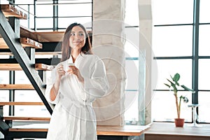 Beautiful Asian woman in white robe holding cup of coffee, standing against the backdrop of staircases