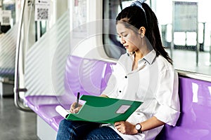 Beautiful asian woman working urgently writing on sheet of paper in a clipboard with pen at the electric train in the morning