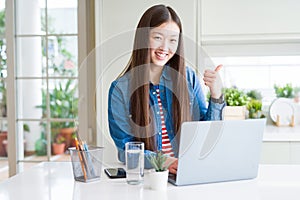 Beautiful Asian woman working using computer laptop happy with big smile doing ok sign, thumb up with fingers, excellent sign