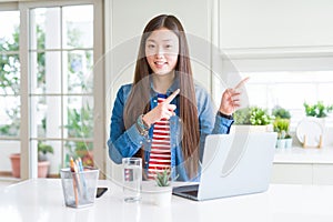 Beautiful Asian woman working using computer laptop smiling and looking at the camera pointing with two hands and fingers to the