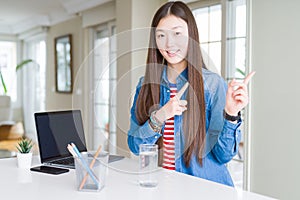 Beautiful Asian woman working using computer laptop smiling and looking at the camera pointing with two hands and fingers to the