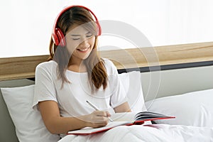 Beautiful asian woman writing diary on the bed in bedroom. She is siting on white bedding.