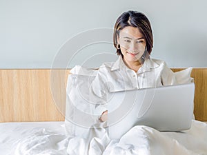 Beautiful Asian women with short hair wearing white shirt working with laptop computer on the bed in the house
