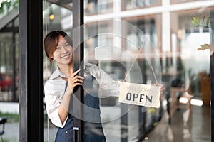 Beautiful asian young barista woman in apron holding tablet and standing in front of the door of cafe with open sign