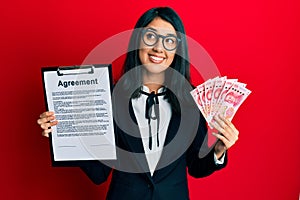 Beautiful asian young business woman holding agreement document and 100 yuan banknotes smiling looking to the side and staring