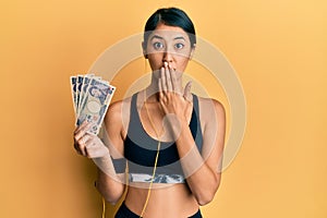 Beautiful asian young sport woman holding 5000 japanese yen banknotes covering mouth with hand, shocked and afraid for mistake