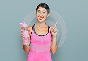 Beautiful asian young sport woman wearing sportswear drinking bottle of water surprised with an idea or question pointing finger