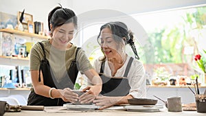 Beautiful Asian young woman enjoy moulding clay making a ceramic plate with aged female