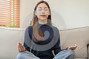 Beautiful asian young woman, girl hands in calm pose sitting practice meditating in lotus position on sofa at home, meditation,