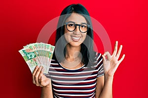 Beautiful asian young woman holding 50 hong kong dollars banknotes doing ok sign with fingers, smiling friendly gesturing