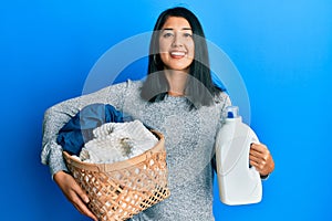 Beautiful asian young woman holding laundry basket and detergent bottle smiling with a happy and cool smile on face