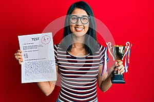 Beautiful asian young woman showing a passed exam holding trophy smiling with a happy and cool smile on face