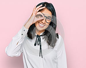 Beautiful asian young woman wearing business shirt and glasses smiling happy doing ok sign with hand on eye looking through
