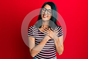 Beautiful asian young woman wearing casual clothes and glasses smiling with hands on chest, eyes closed with grateful gesture on