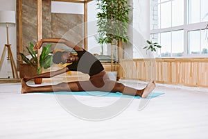 Beautiful and athletic afro american woman sitting and leaning sideways to foot, training on gymnastic mat in decorated