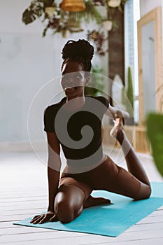 Beautiful and athletic afro american woman sitting and pulling bent rear leg, doing yoga on gymnastic mat in decorated