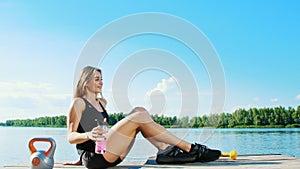 beautiful, athletic blonde girl, woman has rest after training outdoors, drink water. Lake, river, blue sky and forest