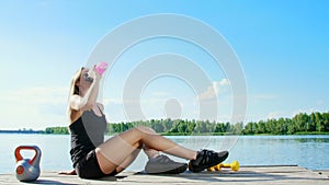Beautiful, athletic blonde girl, woman has rest after training outdoors, drink water. Lake, river, blue sky and forest