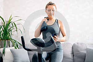 A beautiful, athletic girl with a good mood is engaged in an exercise bike at home and listens to music from her phone through