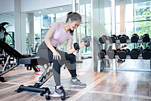 Beautiful athletic muscular Asian woman pumps up the muscles by one arm lifts dumbbell exercise on bench in fitness gym