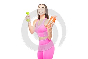 A beautiful, athletic, slim, smiling and cheerful woman in a pink tracksuit holds a dumbbell and a shaker with water