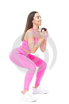 A beautiful, athletic, slim, smiling and cheerful woman in a pink tracksuit performs squats with a pink kettlebell