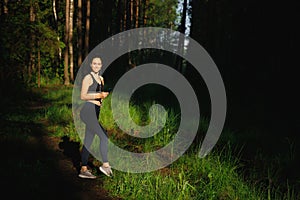 A beautiful athletic Woman in leggings and a t-shirt stands in the forest in the bright sunlight. In the hands of a mobile phone.