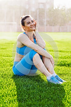 Beautiful, athletic woman with a muscular pumped up body sitting on the grass in urban football stadium. Posing, smiling happily