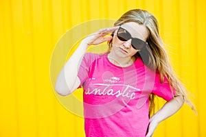 Beautiful athletic young woman with long blond hair in pink shirt, blue jeans. Posing at wall of garage on yellow background.