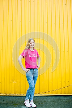 Beautiful athletic young woman with long blond hair in a pink shirt and blue jeans. In white sneakers. Posing and smiling at wall