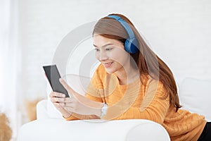 Beautiful Attractive Asian young woman listen music lying on sofa and using smartphone to playing social media in living room